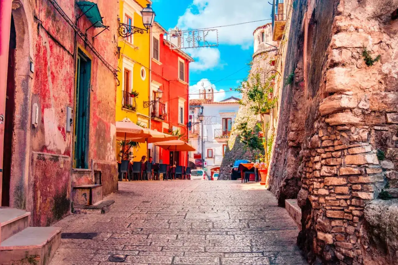 Vico del Gargano, Vicolo del Bacio: un vicolo pittoresco con case colorate, tavolini all'aperto e un'antica muraglia di pietra.