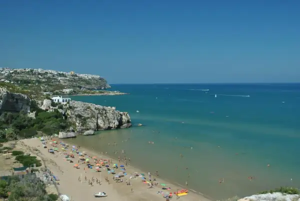 Peschici spiaggia Zaiana, con gente che prende il sole e nuota nelle acque cristalline sotto un cielo azzurro.