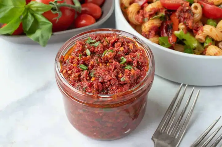 Vaso di pesto di pomodori secchi pugliese, pronto per condire la pasta. Freschi pomodori e basilico sullo sfondo.